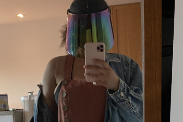 A brown-skinned person wearing a holographic faceshield while taking a mirror selfie.